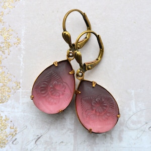 Könnte beinhalten: Ein Paar Ohrringe im Vintage-Stil mit Tropfenform. Die Ohrringe haben einen Farbverlauf aus Rosa und Dunkelviolett mit einem geprägten Blumendesign. Die Ohrringe haben einen goldfarbenen Metallrahmen und Klappbügelverschlüsse.