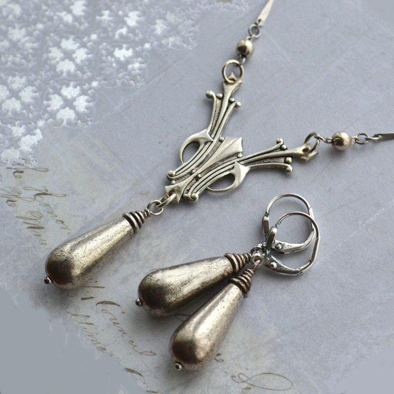 May include: A silver necklace and earrings set with a teardrop pendant and ornate design. The necklace has a chain with small beads and the earrings are drop earrings with a hook closure.