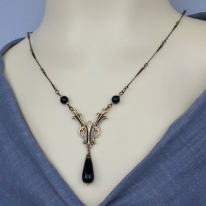 May include: A gold necklace with a black teardrop pendant and black beads. The pendant features an intricate design with a floral motif.