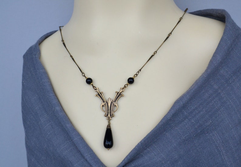 May include: A gold necklace with a black teardrop pendant and black beads. The pendant features an intricate design with a floral motif.