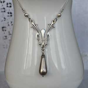 May include: A silver necklace with a teardrop pendant and an intricate design. The necklace is displayed on a white ceramic stand.