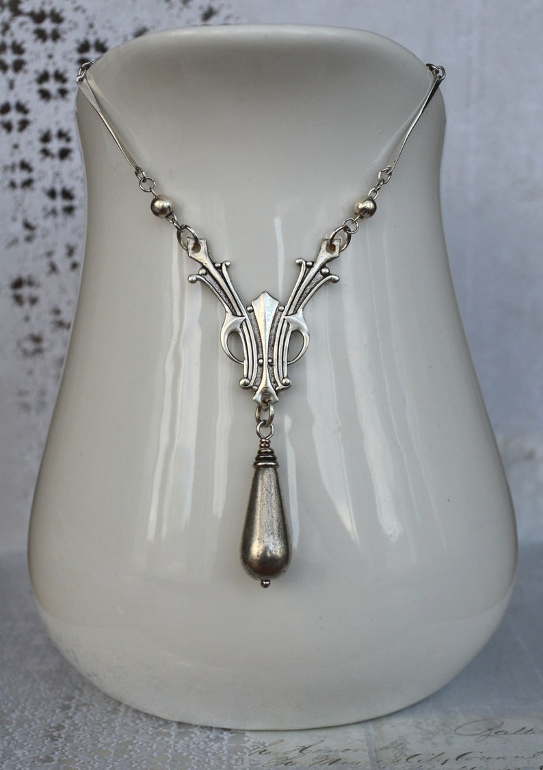 May include: A silver necklace with a teardrop pendant and an intricate design. The necklace is displayed on a white ceramic stand.