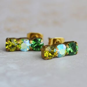 May include: Two pairs of gold-coloured stud earrings, each with three gemstones. The gemstones are arranged in a row, with yellow, iridescent, and green hues. The earrings are set against a soft, blurred background.