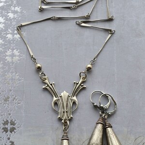 May include: A silver necklace with a pendant featuring a stylized design and two teardrop-shaped earrings. The necklace has a delicate chain and the pendant is detailed with intricate patterns.