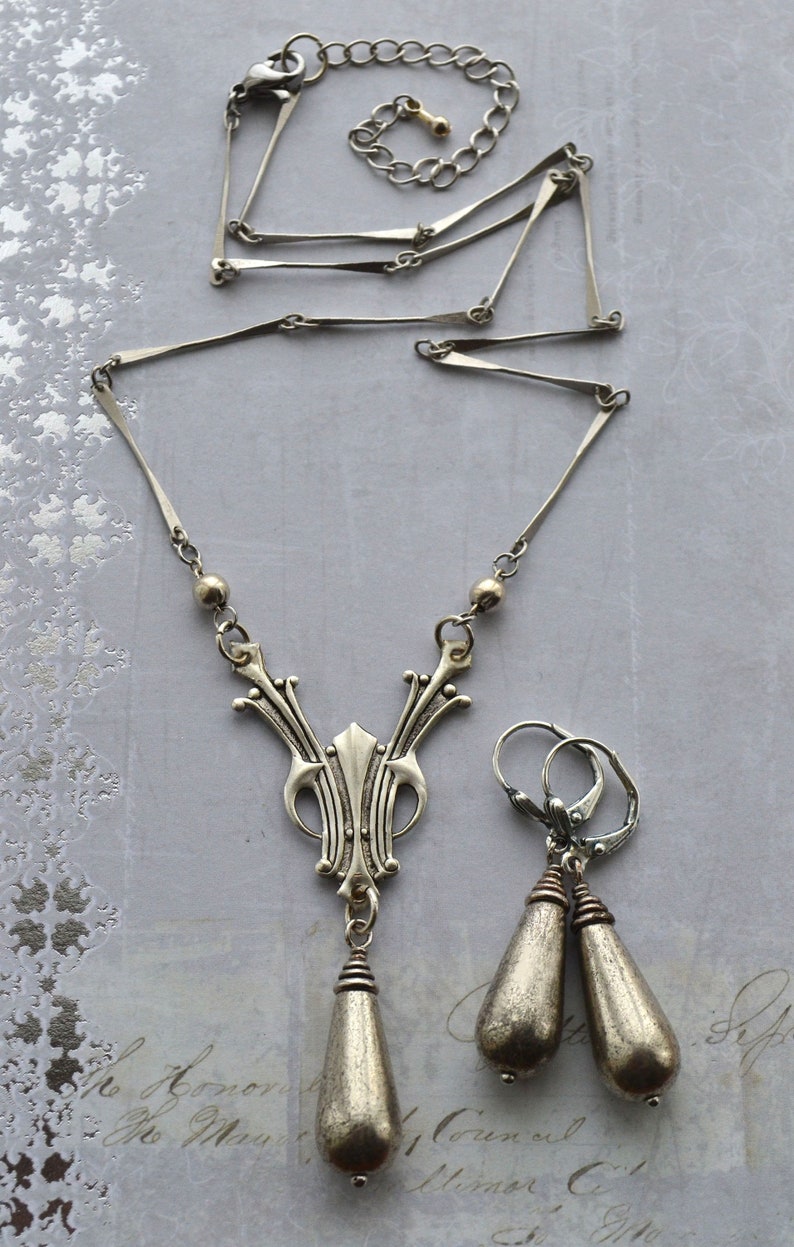 May include: A silver necklace with a pendant featuring a stylized design and two teardrop-shaped earrings. The necklace has a delicate chain and the pendant is detailed with intricate patterns.