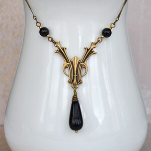 May include: A gold necklace with a black teardrop pendant and black beads. The necklace is displayed on a white ceramic stand.