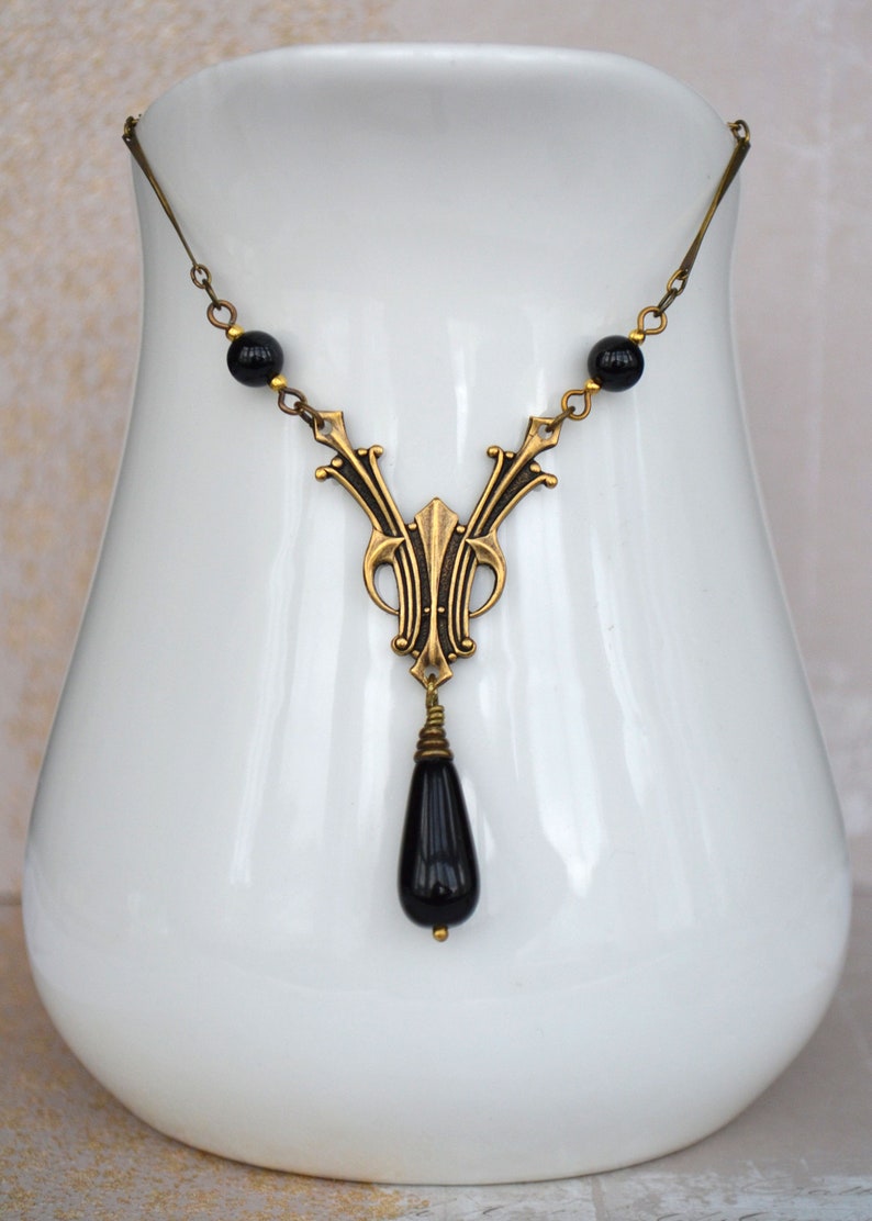 May include: A gold necklace with a black teardrop pendant and black beads. The necklace is displayed on a white ceramic stand.