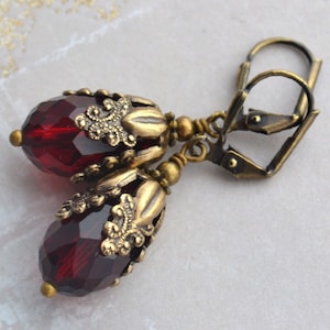 May include: A pair of earrings with a dark red faceted glass teardrop shape set in an ornate gold-toned metal frame. The earrings have a hook closure.