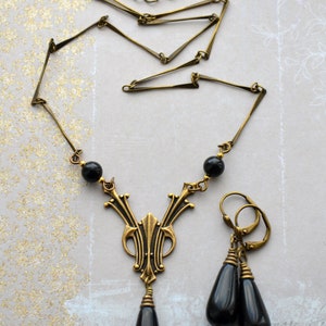 May include: A gold necklace with a black teardrop pendant and matching earrings. The necklace features a geometric design and black beads.