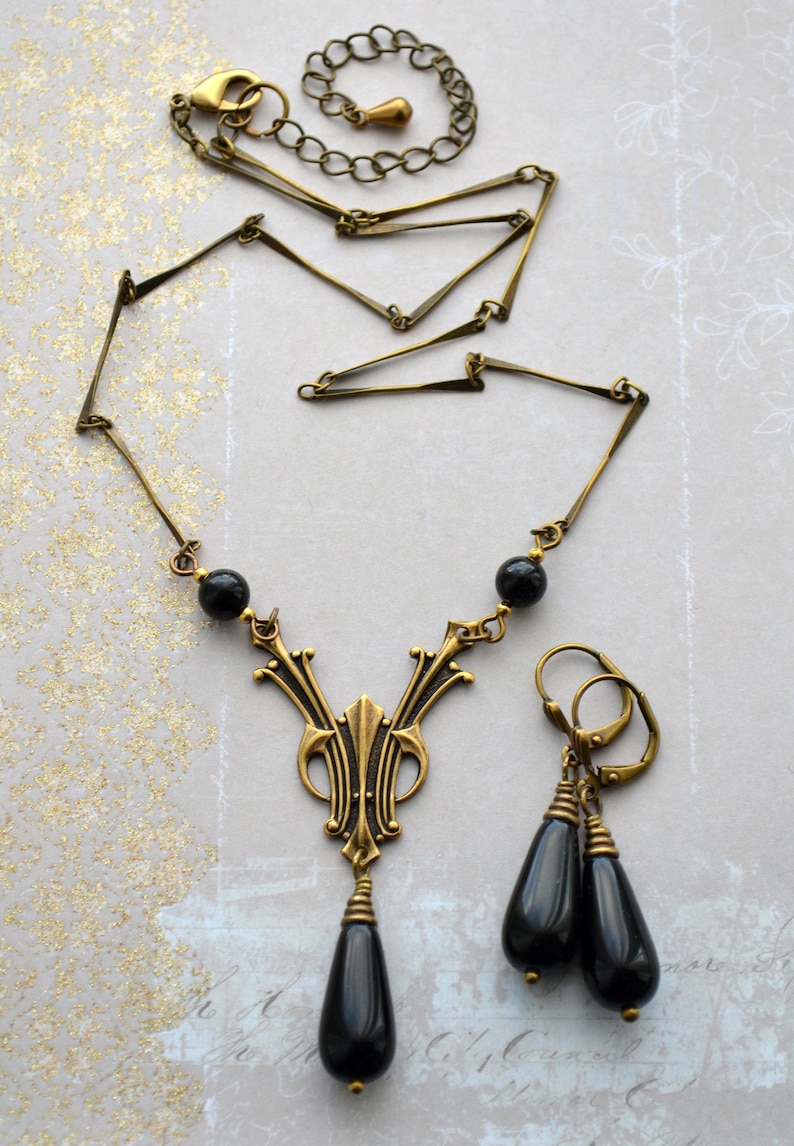 May include: A gold necklace with a black teardrop pendant and matching earrings. The necklace features a geometric design and black beads.