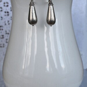 May include: A pair of silver teardrop earrings with a textured finish. The earrings are hanging from a white display stand.