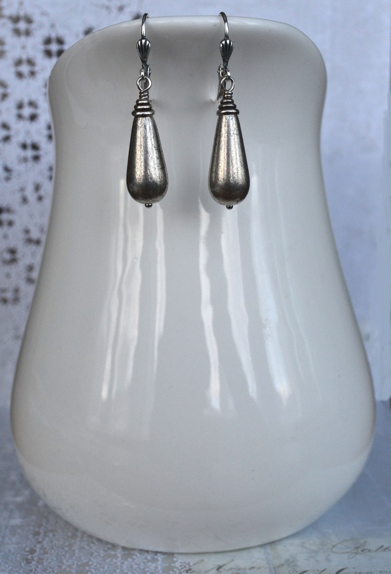 May include: A pair of silver teardrop earrings with a textured finish. The earrings are hanging from a white display stand.