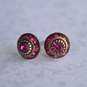 Può includere: Un paio di piccoli orecchini rotondi con una montatura dorata e un centro di strass rosa. Gli orecchini sono circondati da strass rosa più piccoli.