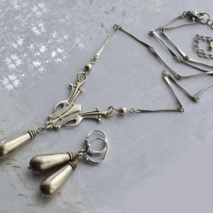 May include: A silver necklace with a geometric pendant and matching earrings. The necklace has a delicate chain with a series of connected bars. The pendant is a stylized design with a pointed top and a rounded bottom. The earrings are simple and dangle from small hoops.