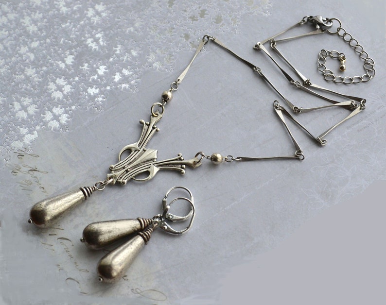 May include: A silver necklace with a geometric pendant and matching earrings. The necklace has a delicate chain with a series of connected bars. The pendant is a stylized design with a pointed top and a rounded bottom. The earrings are simple and dangle from small hoops.