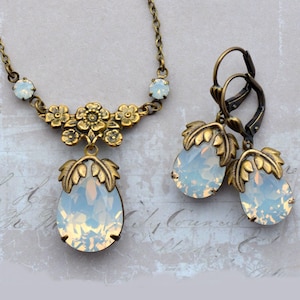 Op de afbeelding: Een vintage-stijl sieradenset met een ketting en bijpassende oorbellen. De set heeft een antiek gouden afwerking met opalescerende, druppelvormige stenen. De ketting heeft bloemen- en bladeraccenten, de oorbellen een vergelijkbaar bladontwerp.