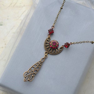 May include: A gold necklace with a pendant featuring a filigree design and a large red rhinestone. The necklace is on a light blue background.