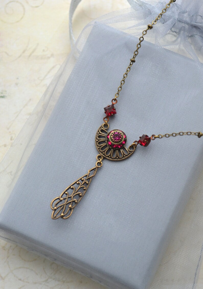 May include: A gold necklace with a pendant featuring a filigree design and a large red rhinestone. The necklace is on a light blue background.