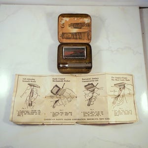 May include: Vintage Ever-Ready safety razor set in a tarnished metal case. The case is open, revealing the razor and a small mirror. A folded instruction sheet with illustrations lies below. The razor is silver-toned.