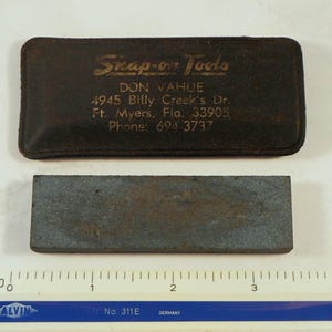 May include: A rectangular, dark gray sharpening stone with a worn appearance, positioned below a dark brown leather case. The case is embossed with "Snap-on Tools" and contact information.