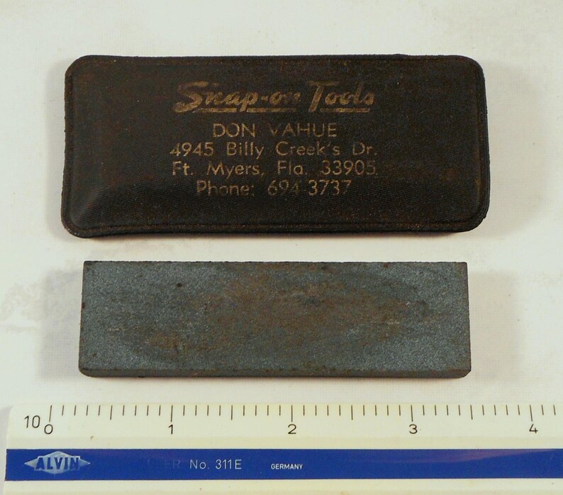 May include: A rectangular, dark gray sharpening stone with a worn appearance, positioned below a dark brown leather case. The case is embossed with "Snap-on Tools" and contact information.