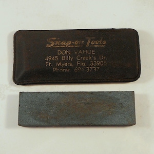 May include: A dark brown leather case with the text "Snap-on Tools" in gold lettering and a gray sharpening stone. The case has the text "DON VAHUE" and an address. The sharpening stone is rectangular.