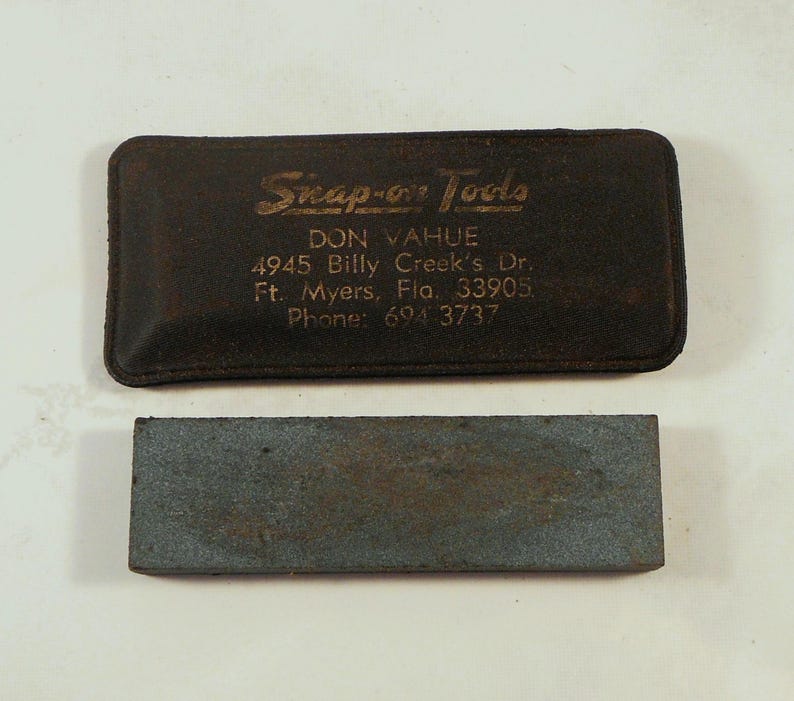 May include: A dark brown leather case with the text "Snap-on Tools" in gold lettering and a gray sharpening stone. The case has the text "DON VAHUE" and an address. The sharpening stone is rectangular.