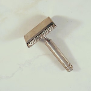 May include: A vintage silver safety razor with a textured handle and a rectangular head. The razor is designed for shaving and has a classic, utilitarian aesthetic. The blade is visible in the head.