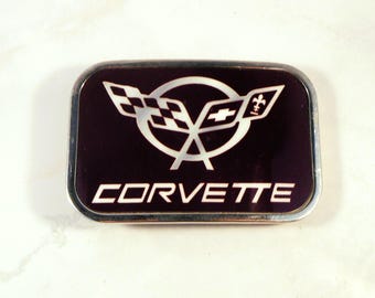 May include: Rectangular emblem with a black background and a silver border. The emblem features the Corvette logo, including crossed flags and the word "CORVETTE" in white capital letters.