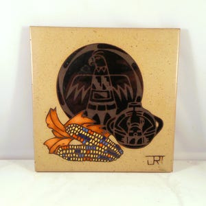 May include: Square ceramic tile with a tan background featuring a Native American design. The design includes a black silhouette of a bird, a pot, and two ears of corn with orange husks and colorful kernels. The tile has a slightly speckled texture.