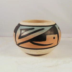 May include: A hand-painted, round ceramic bowl with a cream-coloured base. The bowl features a geometric design in black, light blue, and tan. The rim is a natural beige colour. The design is reminiscent of Native American pottery.