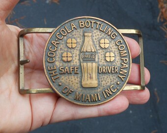 May include: A vintage Coca-Cola Bottling Company belt buckle. The circular bronze buckle features a Coca-Cola bottle and the words "SAFE DRIVER" and "OF MIAMI INC." The buckle has a rectangular brass frame.