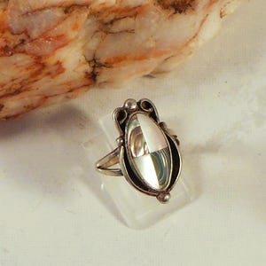 Può includere: Un anello in argento con una montatura ovale allungata caratterizzata da un intarsio segmentato in conchiglia di abalone iridescente e madreperla bianca. La fascia in metallo ha una finitura lucida con piccole sfere decorative in argento.