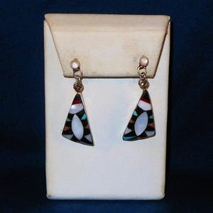 May include: A pair of silver earrings with a triangular design. Each earring features a central white oval surrounded by black, red, and turquoise mosaic-style inlays. The earrings are displayed on a white stand against a dark blue background.