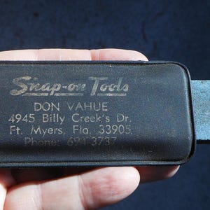 May include: A black Snap-on Tools tool case with a rectangular sharpening stone attached. The case has the text "Snap-on Tools" and address information printed on it. The case is held in a hand.
