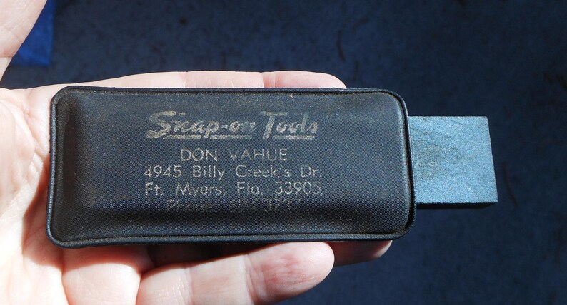 May include: A black Snap-on Tools tool case with a rectangular sharpening stone attached. The case has the text "Snap-on Tools" and address information printed on it. The case is held in a hand.