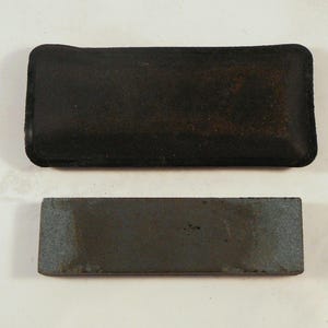 May include: A black rectangular sharpening stone with a dark, textured surface, paired with a black, rectangular case. The stone is gray with darker areas, suggesting use. The case has a slightly worn appearance.
