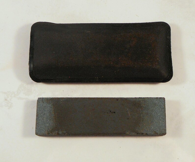 May include: A black rectangular sharpening stone with a dark, textured surface, paired with a black, rectangular case. The stone is gray with darker areas, suggesting use. The case has a slightly worn appearance.