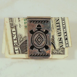 May include: A rectangular silver money clip featuring a black engraved turtle design with a central diamond pattern and wavy line borders, currently holding a folded one-dollar bill.