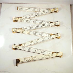 May include: Six vintage white folding rulers with brass-colored joints are arranged on a white surface. The rulers are marked with black numbers and measurement lines. One ruler has the text "LUFKIN" and "DOYLE 18 LOG SCALE 10".