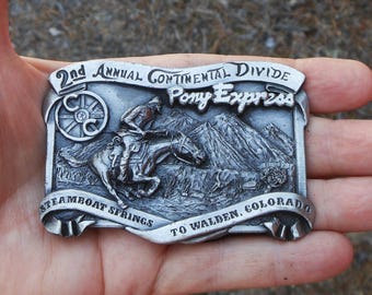 May include: A rectangular silver-toned metal belt buckle featuring a relief design of a rider on horseback. The text reads 2nd Annual Continental Divide Pony Express, Steamboat Springs to Walden, Colorado, with a circular C-C logo on the left.