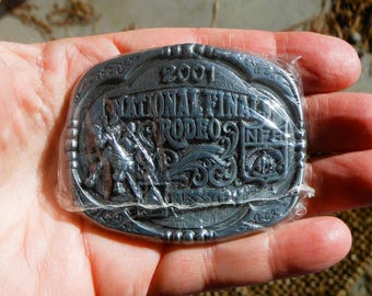 May include: A silver-coloured metal belt buckle from the 2001 National Finals Rodeo. The buckle has an oval shape with a detailed relief design of a rodeo scene and the text "National Finals Rodeo" and "NFR".