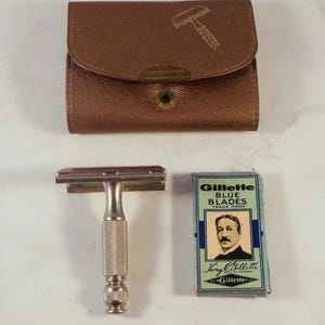Op de afbeelding: Een vintage scheerset met een zilverkleurig veiligheidsscheermes, een bruine leren etui met een scheermesafbeelding en een doosje Gillette Blue Blades. Het scheermes heeft een getextureerde handgreep. De mesjesdoos toont een mannenportret.
