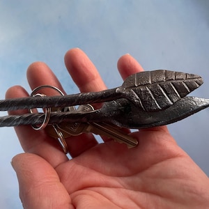 May include: A hand-forged metal key holder with a leaf design. The key holder is dark gray and features a twisted handle and a leaf-shaped end. The keys are attached to the holder with a metal ring.