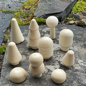 May include: A collection of unfinished wooden decorative objects in various shapes, including cones, trees, spheres, and mushroom-like forms. The objects are arranged on a gray stone surface, showcasing their natural wood grain.
