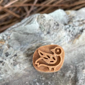 May include: A small, brown, oval-shaped stamp with a carved bird design. The bird has a round eye, a curved wing, and a stylized body. The stamp is likely made of wood or a similar material.