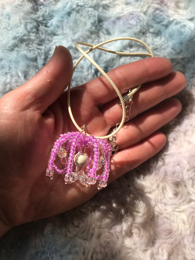 May include: A pink beaded necklace with a white bead pendant. The necklace is on a cream-colored cord.