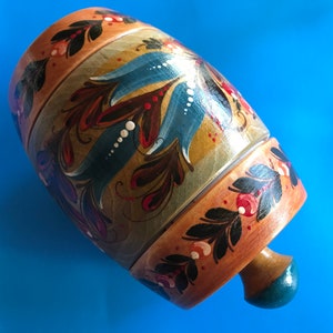 May include: A hand-painted wooden container with a lid and a small handle. The container is cylindrical with a light brown base and is decorated with blue, red, and white floral designs. The background is a solid blue.