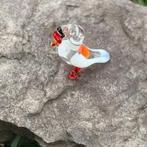 May include: A small glass bird figurine with white, orange, and red accents. The bird is perched on a gray rock.
