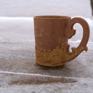 May include: A wooden mug with a carved birch bark design and a decorative handle. The mug is brown and has a light brown and yellow pattern around the top.